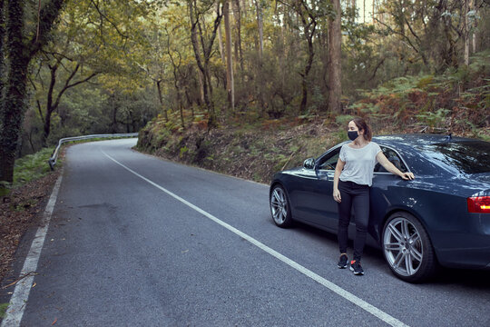 Girl with a mask leaning on a car on a road in the middle of a forest.