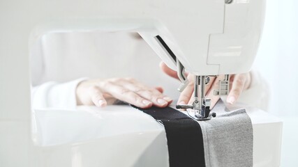 Female hands sew on a white sewing machine close-up. Concept of sewing in modern bright studio, woman in white sweater sewing grey cloth in process