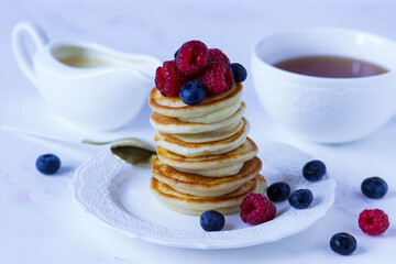 A stack of pancakes with raspberries and blueberries on a white plate against the background of a cup of tea and a gravy boat with condensed milk