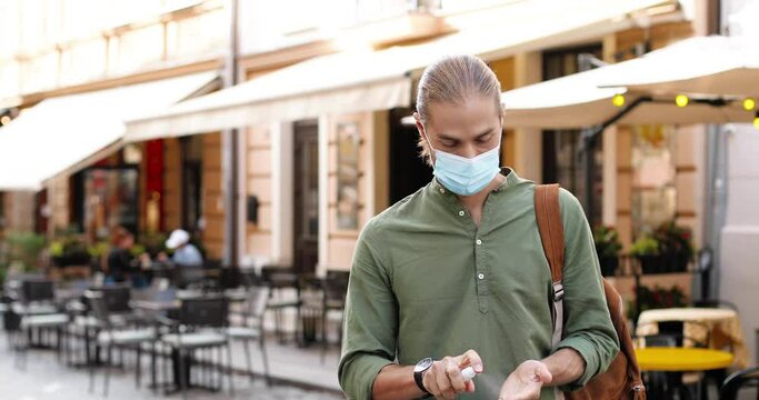Portrait of handsome Caucasian guy in medical mask using hands sanitizer while walking on street in city. Young male in protection sanitizing hands with disinfector outdoor Protective measures concept