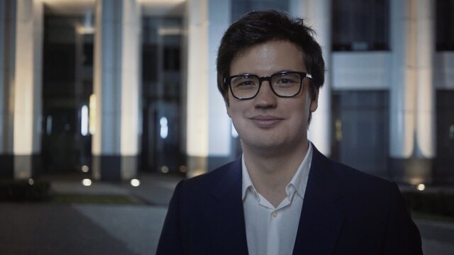Smiling businessman in eyeglasses and black suit at night outside, on background of blue building. Night city shot of man, looking straight at the camera