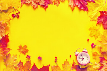 Autumn bright background frame. Yellow-red autumn maple leaves and pink alarm clock on a yellow background, top view, copy space, flat lay. 