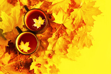 Two cups of tea decorated with a maple leaf and autumn bright yellow-red foliage on a yellow background, top view, copy space. Autumn background