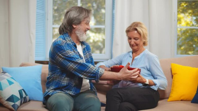 Mature husband presenting packing gift for wife sitting together on couch at home