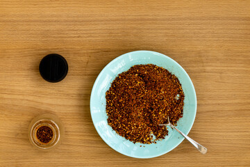 Transferring homemade crushed chili flakes from turquoise color plate to glass spices container. Flat lay view, wooden table. Organic roasted dry chili powder.