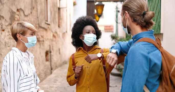 Close up portrait of African American girl meeting with Caucasian female and male friends in medical masks and greeting with elbows outdoor. Multi-ethnic people chatting while standing in town.