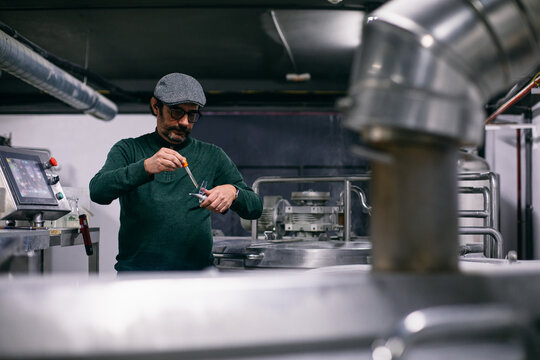 Bearded brewer putting beer sample into refractometer