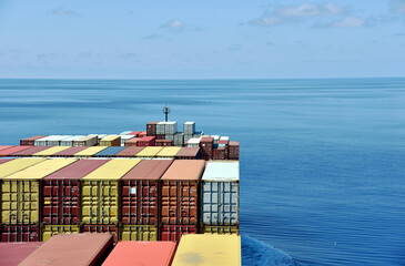 Fully loaded container ship sailing through a blue, calm ocean. View on the stack of the containers loaded on deck of the cargo ship.