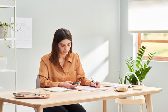 Young businesswoman browsing smartphone while making notes at home