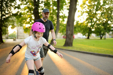 Father teaching his young daughter to roller skate on beautiful summer day in a park. Child wearing...