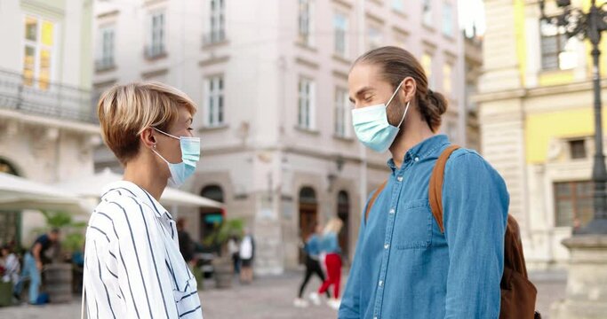 Portrait of Caucasian happy male and female in medical mask greeting with elbows and chatting while standing on street. Handsome young cheerful man speaking with beautiful woman outdoor. Talk concept