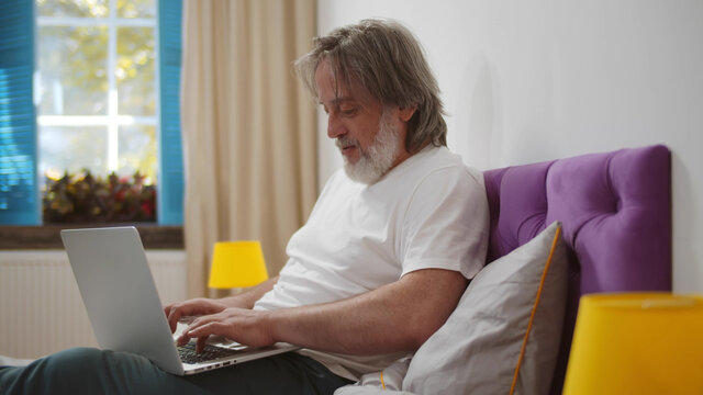Senior man sitting on bed working online from home with laptop