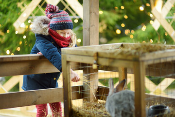 Cute young girl having fun feeding bunnies and sheep in a small petting zoo on traditional Christmas market in Riga, Latvia. Happy winter activities for kids.