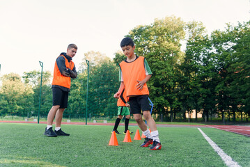 Concentrated junior soccer player running among orange cones that standing on artificial turf at...