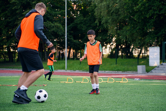 Skillful teen player in football uniform working out the kicking ball together with his experienced coach on sport field