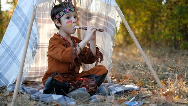 A little boy dressed in a Native American costume, sat down by the wigwam and played the flute. Playing with Indians near the fire with feathers on his head.