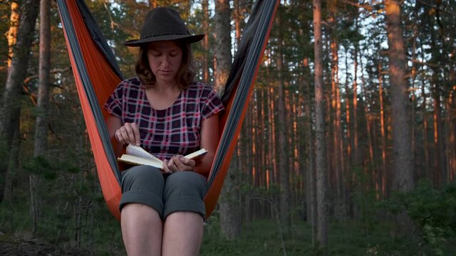 Woman in fedora hat reading book while sitting in hammock in pine forest at sunset. Mindfulness, relax on nature, treat yourself, digital detox, unplugging, life offline concept.