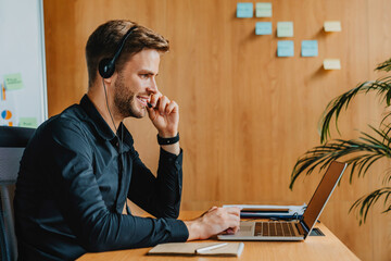 Smiling business man wearing wireless headset make conference video call looking at laptop screen