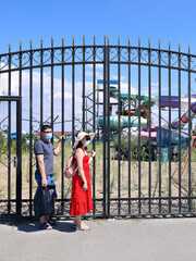 Young man and woman in face masks stand sad next to the closed gates of the water park and look to the side. Inside the territory is abandoned and overgrown with grass without maintenance