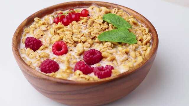 Rotating of wheat flakes porridge with milk, raspberry and currant in wooden bowl on white wooden background and linen textile. Side view, close up.