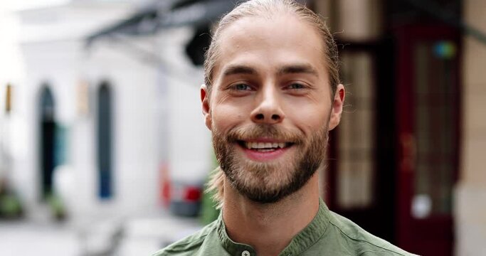 Close up of happy handsome man smiling to camera while standing outdoors. Portrait of cheerful Caucasian bearded guy in good mood looking away on street in city. Positive emotions. Urban concept