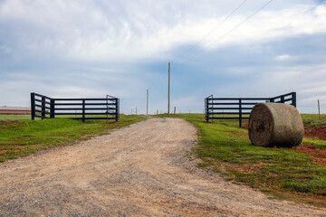 A gravel driveway leads past a round hay bale through an open gate up a gently rolling hill to the...