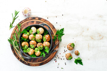 Baked snails with garlic butter and fresh herbs on a black plate on a white wooden background. Top...
