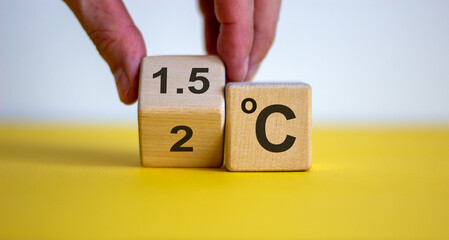 Symbol for limiting global warming. Male hand turnee a cube and changes the expression '2 C' to '1.5 C', or vice versa. Concept. Beautiful yellow table, white background, copy space.