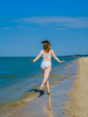 Rear view of a girl running away into the distance along a sandy sea beach Splashes of sea water. The girl is wearing a white swimsuit and sunglasses. Freedom. Fitness. Summer sunny day. Copy space