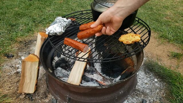 Placing pieces of sausages onto grill to be cooked over fire pit next to cast iron pot of soup.