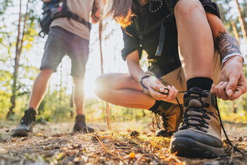 Close up of young female hiker tying shoelaces and getting ready for trekking in forest with man on background. - obrazy, fototapety, plakaty