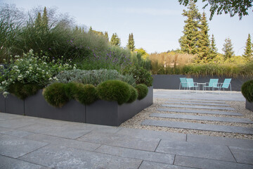Moderne Garten- und Terrassengestaltung im Materialmix: Terrasse und Gehweg aus Naturstein Platten umgeben von Schotter und Metallkübeln mit Grünpflanzen und Ziergräsern im herbstlichen Abendlicht