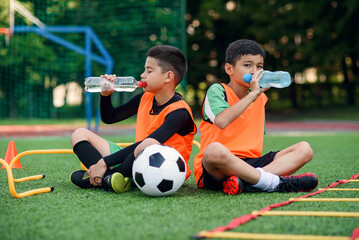 Two teen boys in football uniforms having a rest on sport field with artificial covering and...