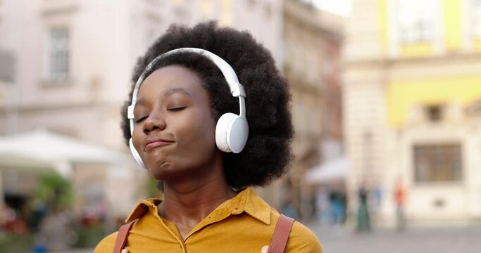 Caucasian happy beautiful African American female in headphones walking on street and listening to music outdoors. Joyful girl in good mood enjoying songs in town. Close up portrait. Leisure concept