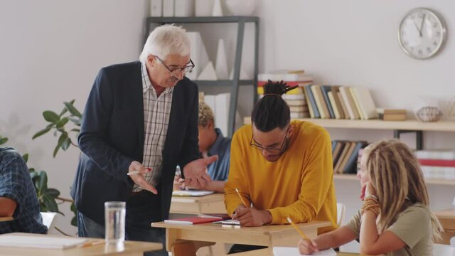 Senior professor explaining something to young male student of mixed-race ethnicity as he taking notes at desk in college classroom