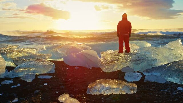 Man stands on iceberg on Okulsarlon Beach at sunset, slow motion
