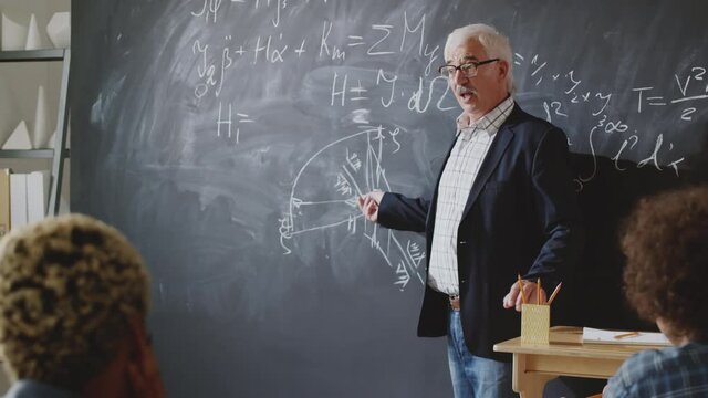Senior professor explaining graph on chalkboard during math lesson, then looking at camera and smiling while giving lesson to college students
