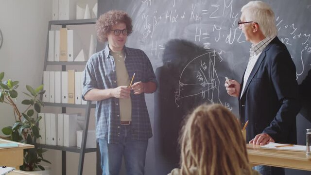 Young male student pointing at formulas written on chalkboard and speaking to senior professor and groupmates while reciting a lesson in high school