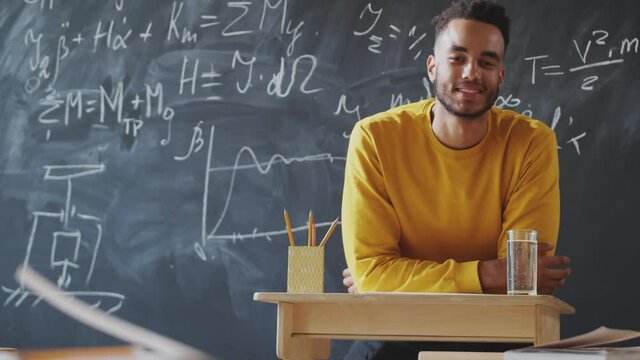 Young handsome male student of mixed-race ethnicity looking at camera and smiling while leaning on lectern beside chalkboard in college classroom