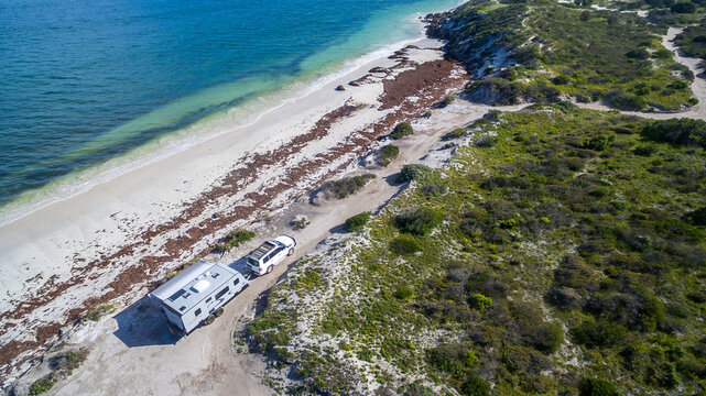 Landscape aerial view of 4WD and modern caravan parked adjacent to a sparkling sunny beach.
