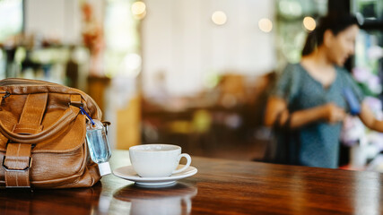 Mini alcohol gel bottle to kill Corona Virus(Covid-19) hang on a brown leather shoulder bag with hot coffee on table in cafe.New normal lifestyle.Health care concept.Selective focus on alcohol gel