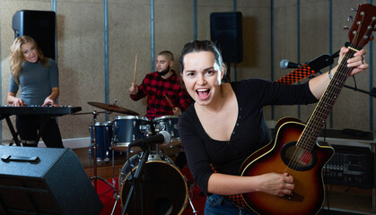 Fototapeta na wymiar Attractive happy female soloist playing guitar and singing with her music band in sound studio