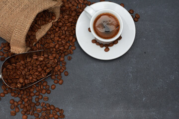 White espresso cup with foam, coffee beans in scoop, burlap sack on dark table background with copy space. Arabica grains. Coffee shop, caffeine, roast concept
