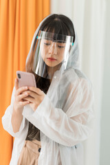 Asian woman wearing white raincoat and face shield holding smartphone.