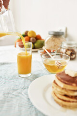 The process of pouring orange juice in the glass, Honey in a bowl with dipper, pancakes and various fruits served for breakfast on the table