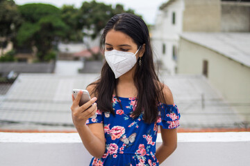 Ni&ntilde;a latina con protecci&oacute;n mascarilla con celular desde su balc&oacute;n