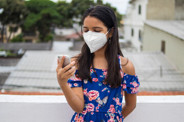 Ni&ntilde;a latina con protecci&oacute;n mascarilla con celular desde su balc&oacute;n