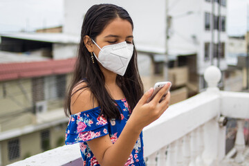Ni&ntilde;a latina con protecci&oacute;n mascarilla con celular desde su balc&oacute;n