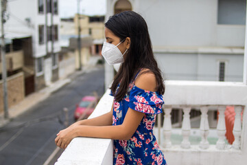 Ni&ntilde;a latina con protecci&oacute;n mascarilla pensando desde su balc&oacute;n