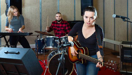 Fototapeta na wymiar Attractive positive female soloist playing guitar and singing with her music band in sound studio